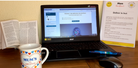Un ordinateur portable affiche un cours en ligne, avec un livre ouvert, un guide imprimé, un stylo, une souris et un mug étiqueté "Mum's the Greatest" (Maman est la meilleure) disposés sur un bureau.