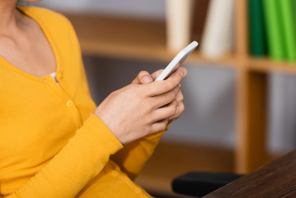 Personne en pull jaune tenant et utilisant un smartphone, assise à l'intérieur près d'un bureau et d'étagères, consultant peut-être le Bonus du livre sur son appareil.