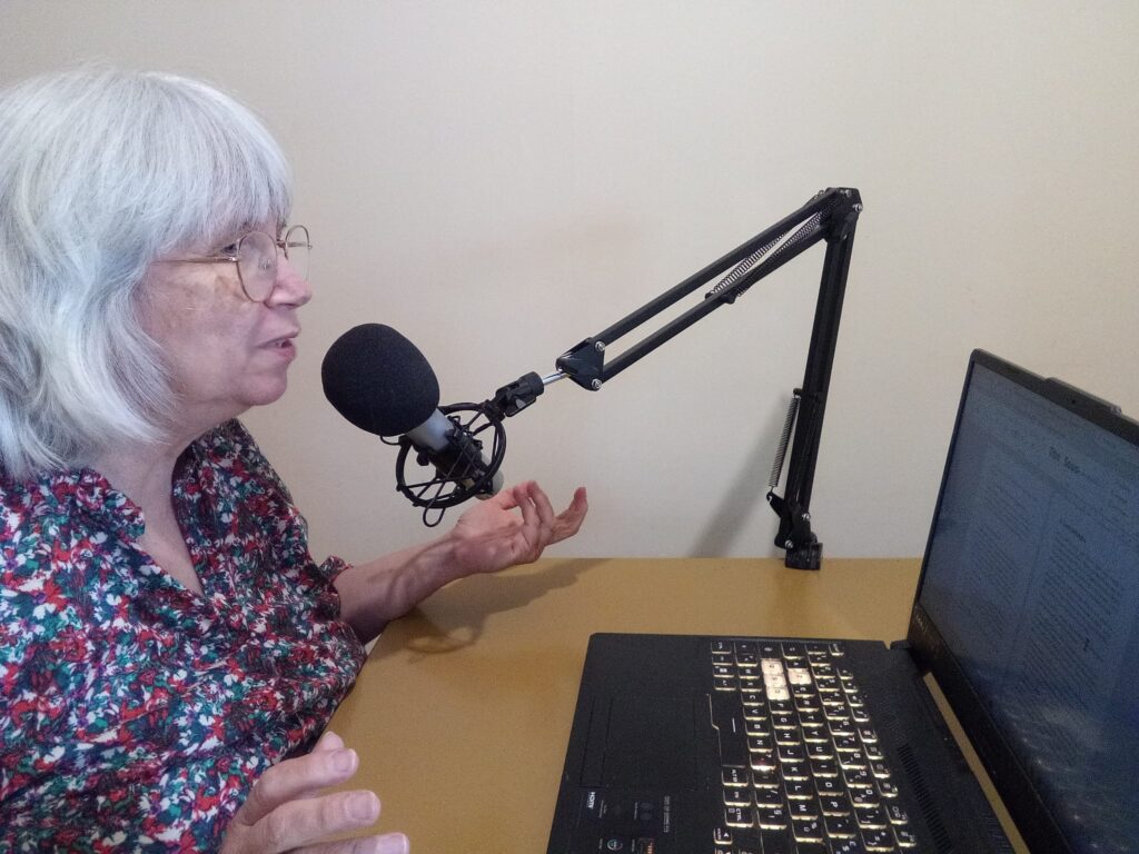 Une femme âgée aux cheveux grisonnants parle dans un microphone monté sur une perche, assise à un bureau avec un ordinateur portable ouvert, partageant ses idées sur le bonus du livre.